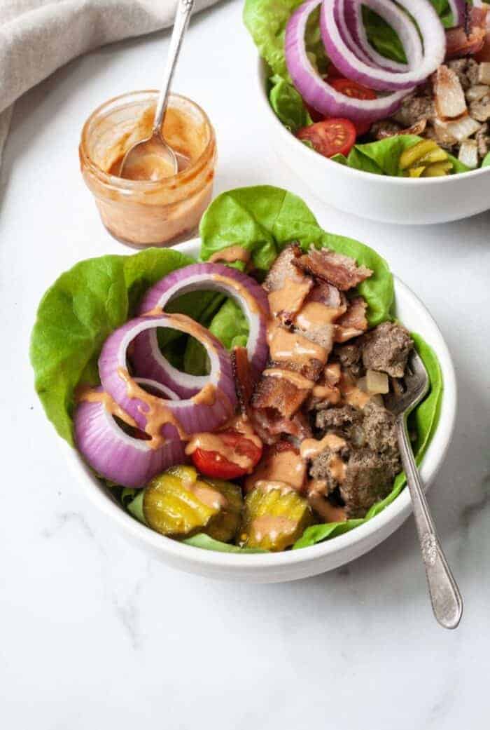 A white bowl filled with ground beef, onions, lettuce, pickles, and tomatoes.