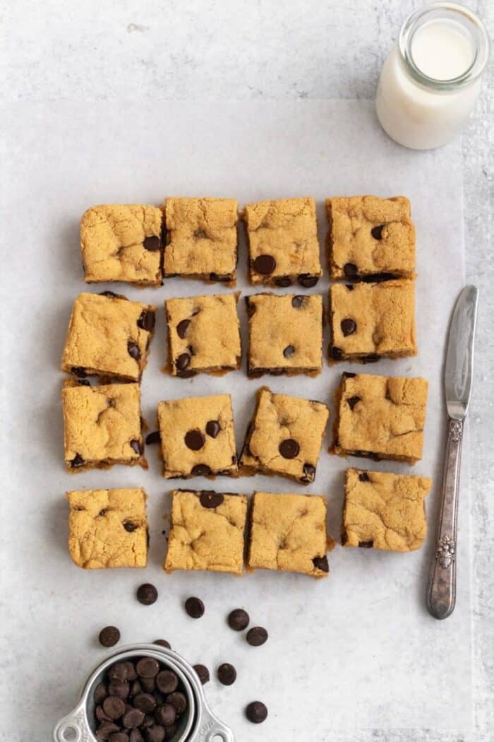Almond flour blondies on a light colored backdrop with chocolate chips and a glass of milk.